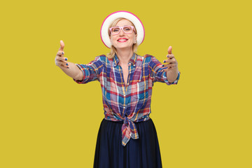 Portrait of happy modern stylish mature woman in casual style with hat and eyeglasses standing giving hands to hug and looking with toothy smile. indoor studio shot isolated on yellow background.