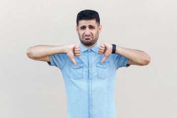 Portrait of sad, upset or worry handsome young bearded man in blue shirt standing, thumbs down and looking at camera with dissatisfied face. indoor studio shot isolated on light beige wall background.