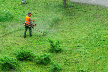 A communal service worker in an orange vest mows grass in a park with a hand gas mower