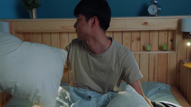 Young tired Asian man trying to sleep on bed in bedroom at night, covering himself with blanket. Noisy neighbors, stressful situation, exhausted man. Bad mood, negative emotions. Male portrait