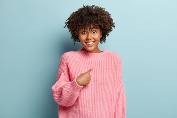 Human reaction concept. Positive dark skinned woman has crisp hair, points at herself, looks gladfully, wears oversized pink jumper, cannot believe she is winner, isolated over blue studio wall - obrazy, fototapety, plakaty