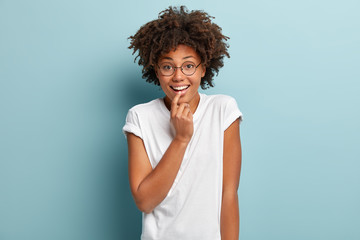 Fototapeta na wymiar Tenderness, good emotions and sincere feelings concept. Portrait of gentle dark skinned woman touches lower lip, has toothy smile, wears round spectacles and casual t shirt, glad hear good news