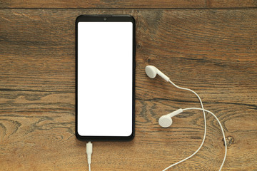 Smartphone with isolated white blank screen with connected earphones on the background of a wooden desk