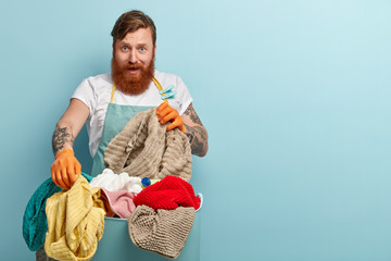 Frustrated redhaired man with trendy haircut, thick beard, holds dirty laundry from basket, wears rubber protective gloves, isolated on blue background with copy space for your advertising content
