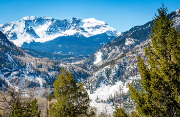 snow capped mountains and blue skies in these wintery scenics of nature as spring breaks through the cold snow of winter to show brown earth and green trees in Colorado