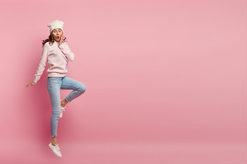 Stupefied young European woman with curly hair, wears stylish clothing, jumps high in air, poses sideways in full length, isolated over pink background with copy space for your promotion or text © WHstudio Leushin N