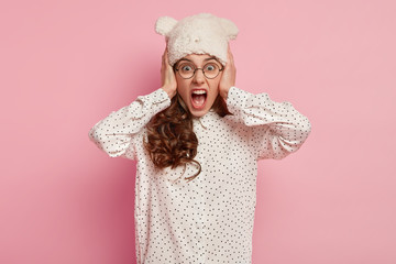 Half length shot of emotional irritated cute woman screams loudly, covers ears, expresses negative emotions, wears warm white hat, polka dot shirt, has curly hair, isolated on pink background