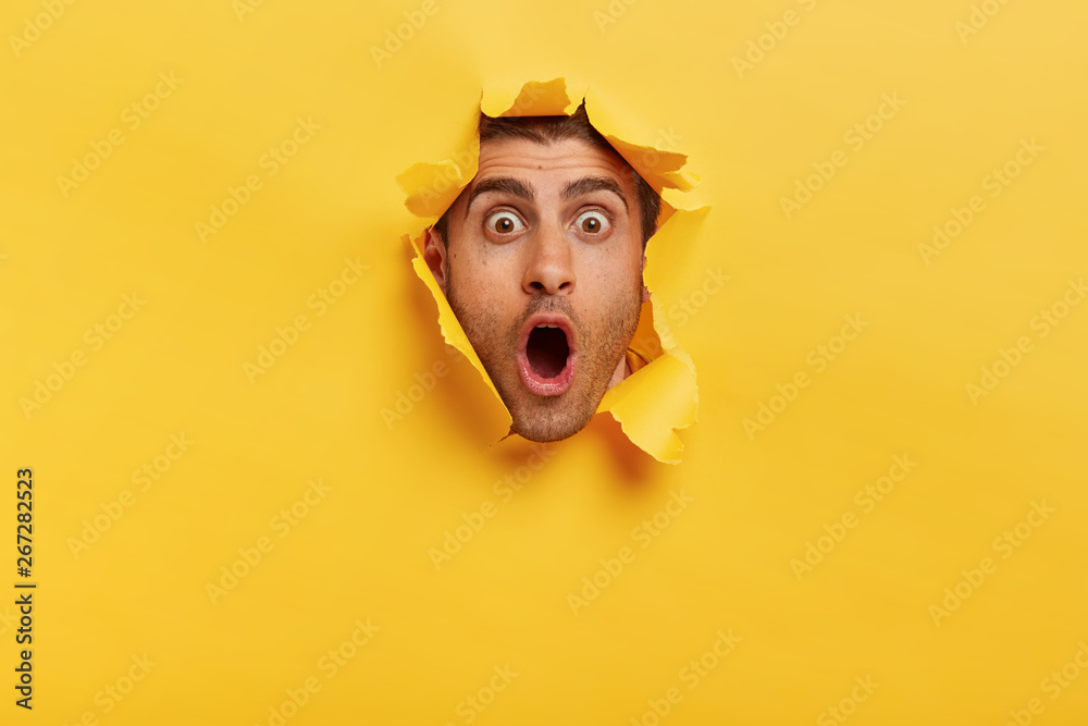 Wall mural Stupefied young man with bristle keeps jaw dropped from surprisement, has popped eyes, stares through yellow torn paper, impressed by shocking rumors. People, human emotions and reaction concept