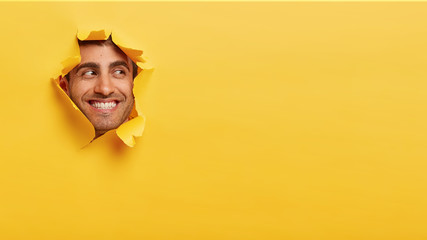 Happy satisfied man with toothy smile, concentrated aside, sees something awesome, looks through hole in colored paper, enjoys photographing in studio. Handsome guy breaks through yellow wall