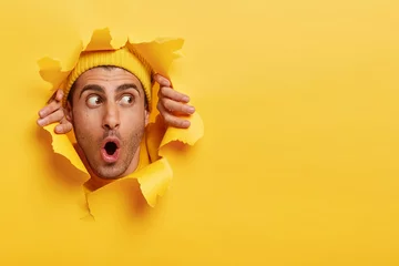 Deurstickers Surprised male face through paper hole. Emotional astonished young man wears yellow headgear, makes slot in background with hands, keeps mouth wide opened. Copy space to insert yout text or slogan © Wayhome Studio
