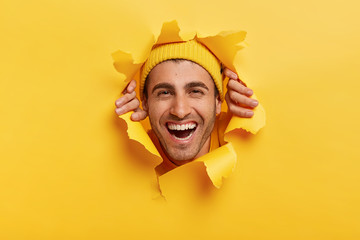 Positive unshaven male adult looks happily through yellow paper, shows only face, wears hat, shows...