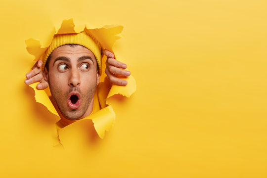 Surprised male face through paper hole. Emotional astonished young man wears yellow headgear, makes slot in background with hands, keeps mouth wide opened. Copy space to insert yout text or slogan