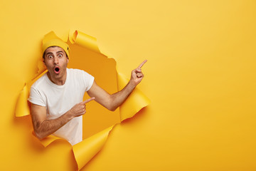 Horizontal shot of surprised young Caucasian man in white t shirt and yellow headgear, has widely opened mouth, indicates at copy space, models in torn paper background. Wow, just look there