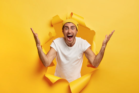 Brilliant! Optimistic overjoyed unshaven European man raises hands, impressed by something awesome, rejoices sale and discounts, wears yellow hat and white casual t shirt, breaks through paper