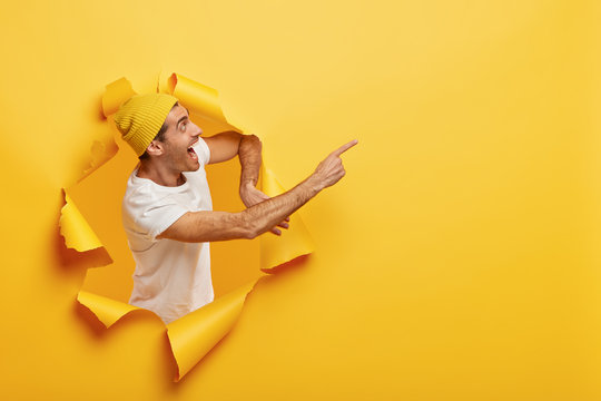 Photo of overjoyed hipster looks gladfully away, points fore finger on blank space, shouts from joy, stands through torn paper hole, wears stylish yellow hat and casual white t shirt. Promotion