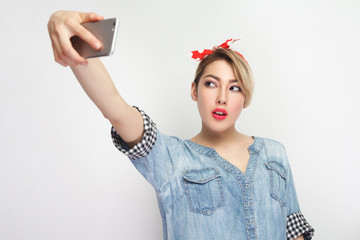 Selfie time! Portrait of happy attractive blogger woman in casual blue denim shirt with makeup, red headband standing, holding phone and making selfie. indoor studio shot, isolated on white background