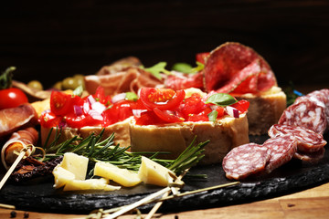 Traditional italian antipasto bruschetta appetizer with cherry tomatoes, cream cheese, basil leaves and balsamic vinegar on cutting board with prosciutto, salami, cheese