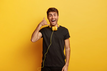 Waist up shot of positive Caucasian man makes call gesture, listens music in headphones happily, dressed in black casual t shirt, isolated over yellow background. Body language concept. Call me back