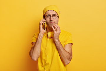 Photo of serious attentive young man has phone conversation, keeps modern cellphone near ear, has headphones, wears yellow clothes, poses indoor, finds out new information. Communication concept