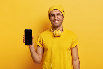 Satisfied European man holds modern cellular with black mockup space, wears yellow headgear, t shirt, has headphones on neck, advertises nice gadget for customers. Technology and people concept