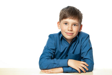 The child sits at the table and dreams of something. Portrait of a child in a blue shirt, sitting behind a desk, isolated, on a white background.