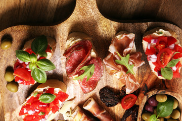 Traditional italian antipasto bruschetta appetizer with cherry tomatoes, cream cheese, basil leaves and balsamic vinegar on cutting board with prosciutto, salami, cheese