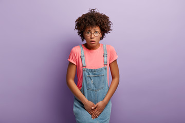 Dissatisfied girl keeps hands on crotch, presses lower abdomen, needs toilet badly, has syndrome of cystitis, wears spectacles, pink t shirt and denim sarafan, isolated on purple wall. Health problem - obrazy, fototapety, plakaty