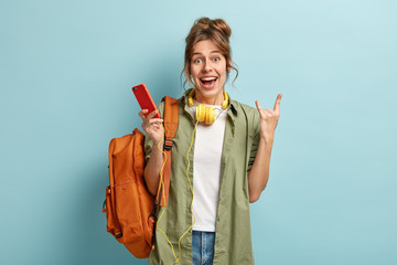 Photo of cheerful Caucasian woman makes horn gesture, holds smart phone, listens audio book, feels...