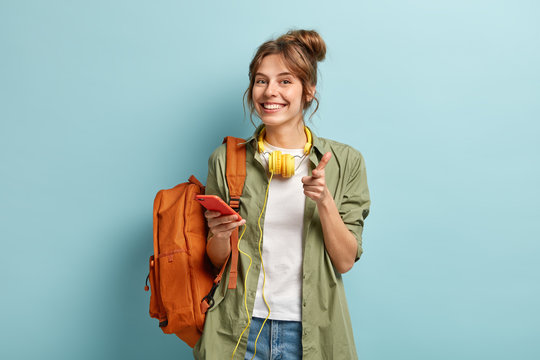 Photo of delighted European woman points at you happily, holds smartphone device connected to headphones, uses music app, smiles gladfully, dressed in long casual shirt, stands with rucksack