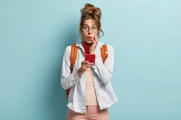 Half length shot of impressed young woman with scared expression, holds modern cell phone, being hitchhiker traveler, searches hotels in city, carries rucksack on back, wears casual shirt and trousers