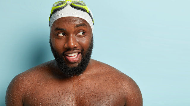 Close up shot of happy thoughtful man focused aside, being wet after going out of water, wears goggles and swimcap, enjoys recreation time, isolated over blue background with copy space aside
