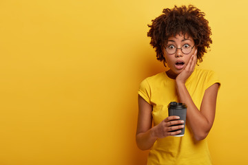 Photo of surprised dark skinned woman looks in stupor at camera, keeps hand on cheek, opens mouth...