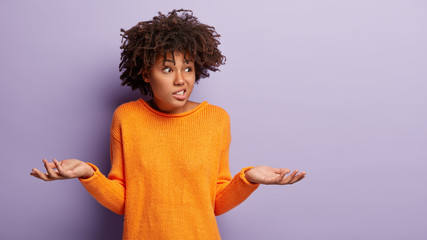 Photo of doubtful Afro woman faces dilemma, spreads hands sideways, clenches teeth, focused aside...