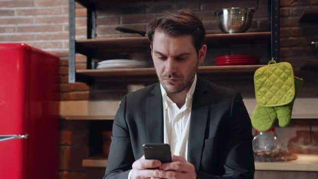 Rotation view of stylish attractive businessman using his mobile phone at home, actively typing messages on modern cozy kitchen background. Looking good, modern gadgets, communication concept. Male