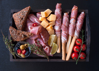 Antipasto is a cold meat dish with grissini breadsticks, prosciutto, ham, ham slices, beef jerky, salami and cheese on a wooden tray on a black wooden background. The view from the top. Copy space