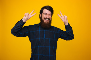 A cheerful man with beard and toothy smile, doing peace gesture over yellow background