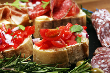 Traditional italian antipasto bruschetta appetizer with cherry tomatoes, cream cheese, basil leaves and balsamic vinegar on cutting board with prosciutto, salami, cheese