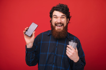 Photo of excited bearded man showing his phone while holding a cup with coffee, red wall