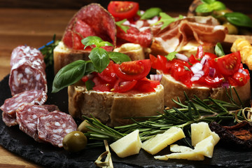 Traditional italian antipasto bruschetta appetizer with cherry tomatoes, cream cheese, basil leaves and balsamic vinegar on cutting board with prosciutto, salami, cheese