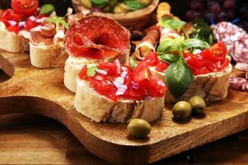 Traditional italian antipasto bruschetta appetizer with cherry tomatoes, cream cheese, basil leaves and balsamic vinegar on cutting board with prosciutto, salami, cheese
