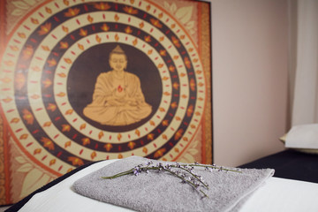 In a healing massage studio on the wall a large East Asian buddha image, in the foreground a massage table, with a white cushion, a towel and a branch of lavender on it