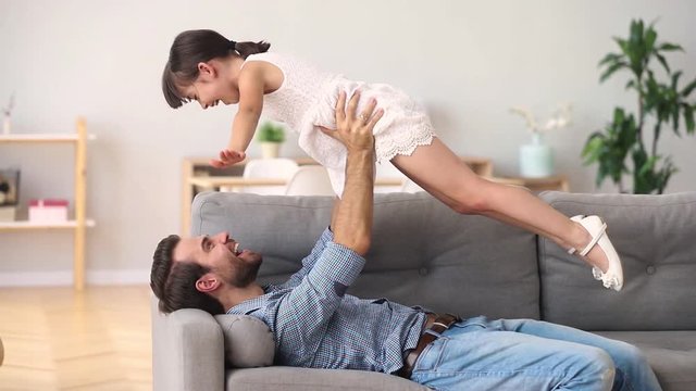 Happy father lying on couch lifting child daughter having fun