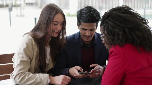 Serious man and attractive Caucasian woman swiping photos on phone, choosing while having meeting with pretty Afro-american woman. Wedding preparation, house searching. Communication concept
