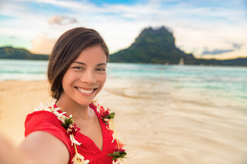 Luxury vacation selfie happy woman smiling taking photo with mobile phone on Bora bora island cruise travel destination at sunset. Smiling gorgeous Asian girl wearing Tahiti flower lei for luau. - obrazy, fototapety, plakaty