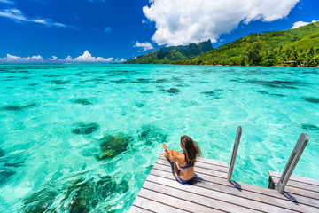 Bora bora luxury travel overwater bungalow resort vacation bikini woman at Tahiti hotel. Tropical exotic destination. Girl relaxing sitting on private balcony under the sun looking at ocean. - obrazy, fototapety, plakaty