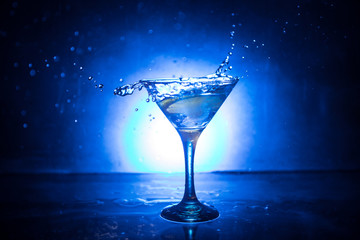 Martini cocktail glass in hand splashing on dark toned smoky background or colorful cocktail in glass with splashes and olives. Party club entertainment. Mixed light. Selective focus