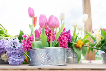 Tin flower pots with a floral arrangement of tulips and other flower varieties in pink, white, yellow and bright green stems. Plywood work table showing.