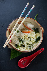 Bowl of noodle soup with wontons on a black stone background, vertical shot, elevated view
