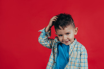 Photo of a charming boy who looks into the camera, smiles and scratches his head with a hand kiss on a red background. Human emotions