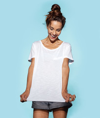 Portrait of brunette girl with tied up hairstyle showing copy space on white shirt. Cheerful model posing on blue background. Positivity and happiness concept